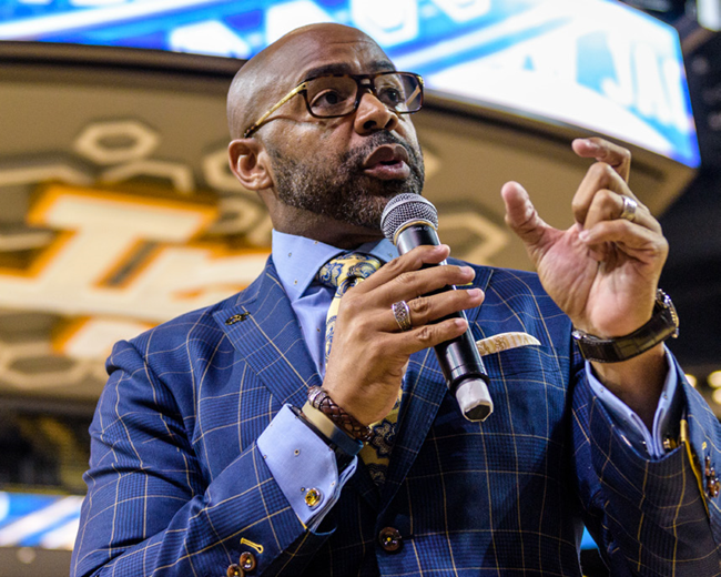 Kofi Smith in blue suit speaking into microphone and gesturing with his hand