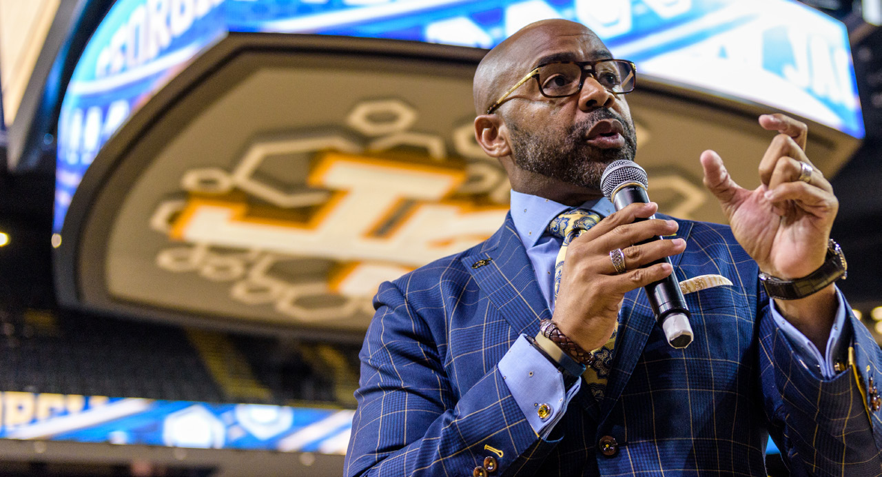 Kofi Smith in blue suit speaking into microphone and gesturing with his hand